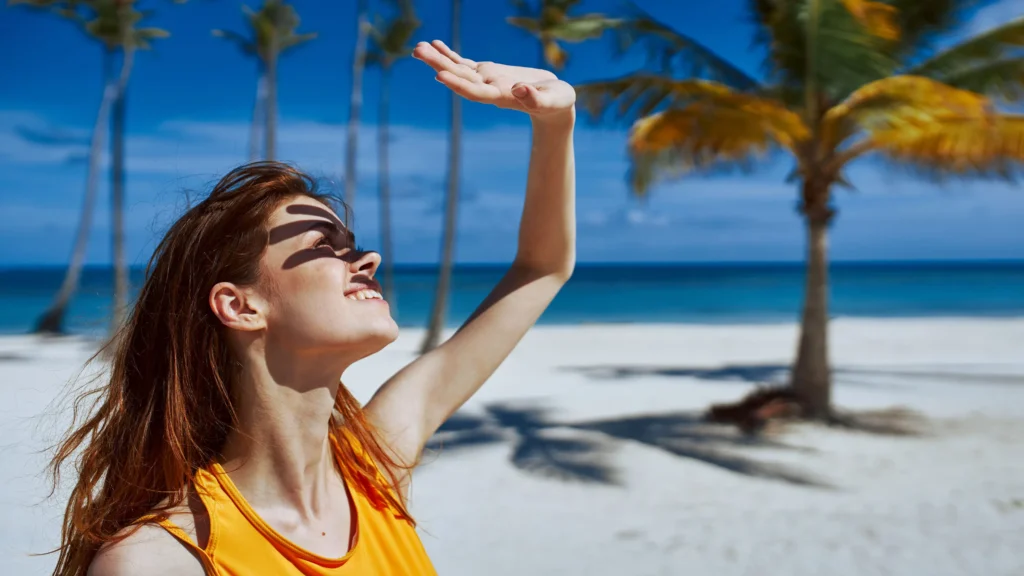 Young woman shielding her face from the sun at a tropical beach, practicing sun protection under clear skies