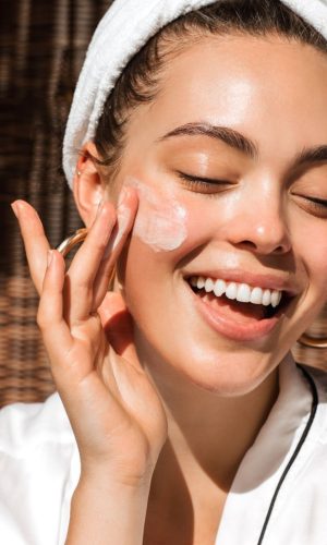 Smiling woman applying face cream to her cheek during skincare routine.